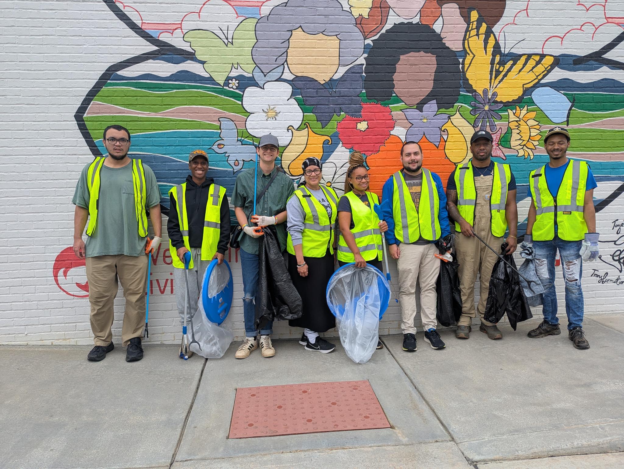 8 people standing in front of a mural with trash pick-up gear.